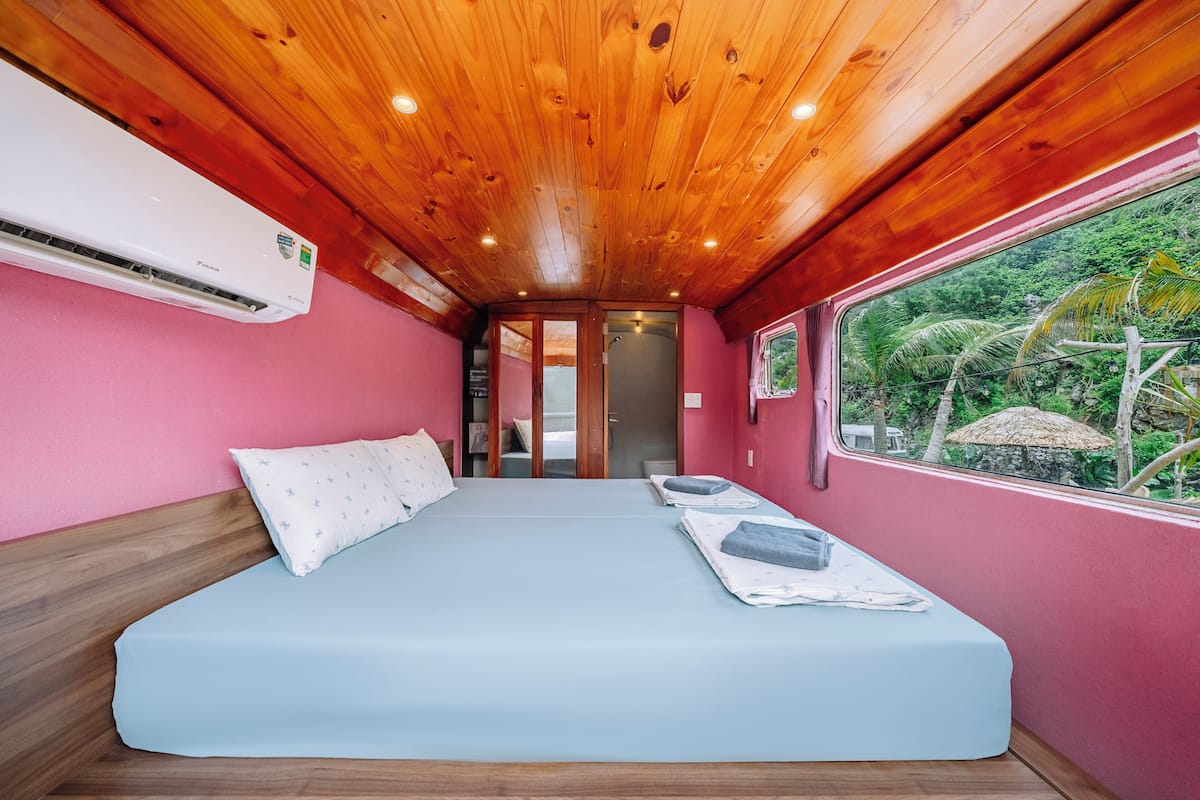 Una habitación con cama, techo de madera, paredes rosas y ventana con vista a follaje.