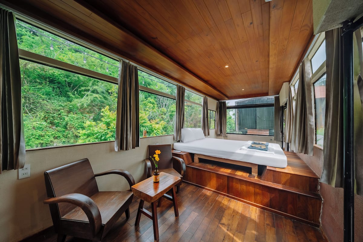 Una habitación con techo de madera, ventanas amplias, una cama, una mesa con un florero y un banco.
