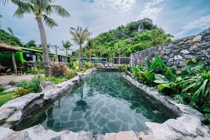 A clear water feature surrounded by greenery and stone walls, with a view of a green bus and a thatched-roof structure in the background. - The Healing Camp - Uncommon Stay (Hai Phong)