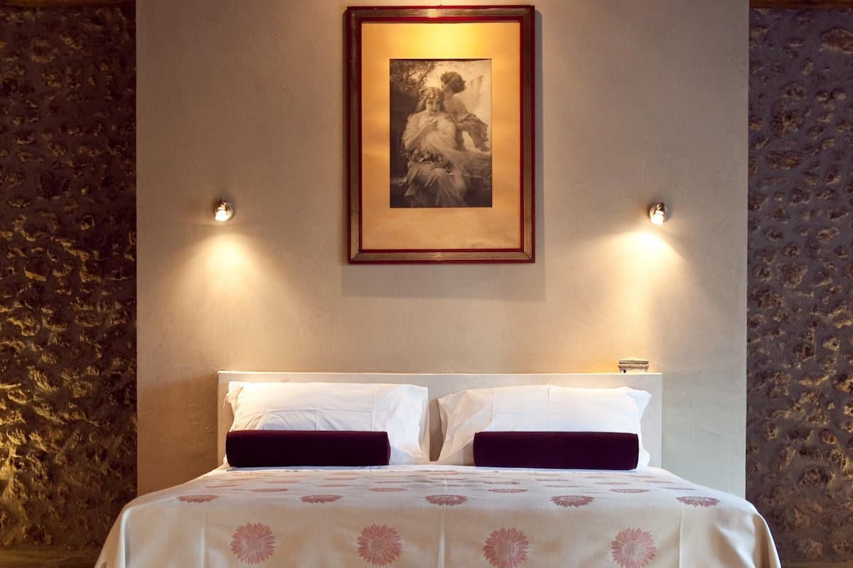 Ein Bett mit weißen Bettwäsche und dunklen Kissen, ein gerahmtes Bild darüber und eine Steinwand links.