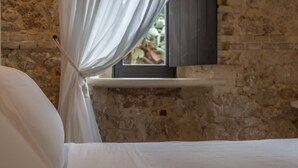 A bed with white linens, a window with a curtain, and a television on a stone wall. - Mandranova Farm (Palma di Montechiaro)