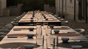 A long dining table set with chairs, glasses, and cutlery, with a building and trees in the background. - Mandranova Farm (Palma di Montechiaro)
