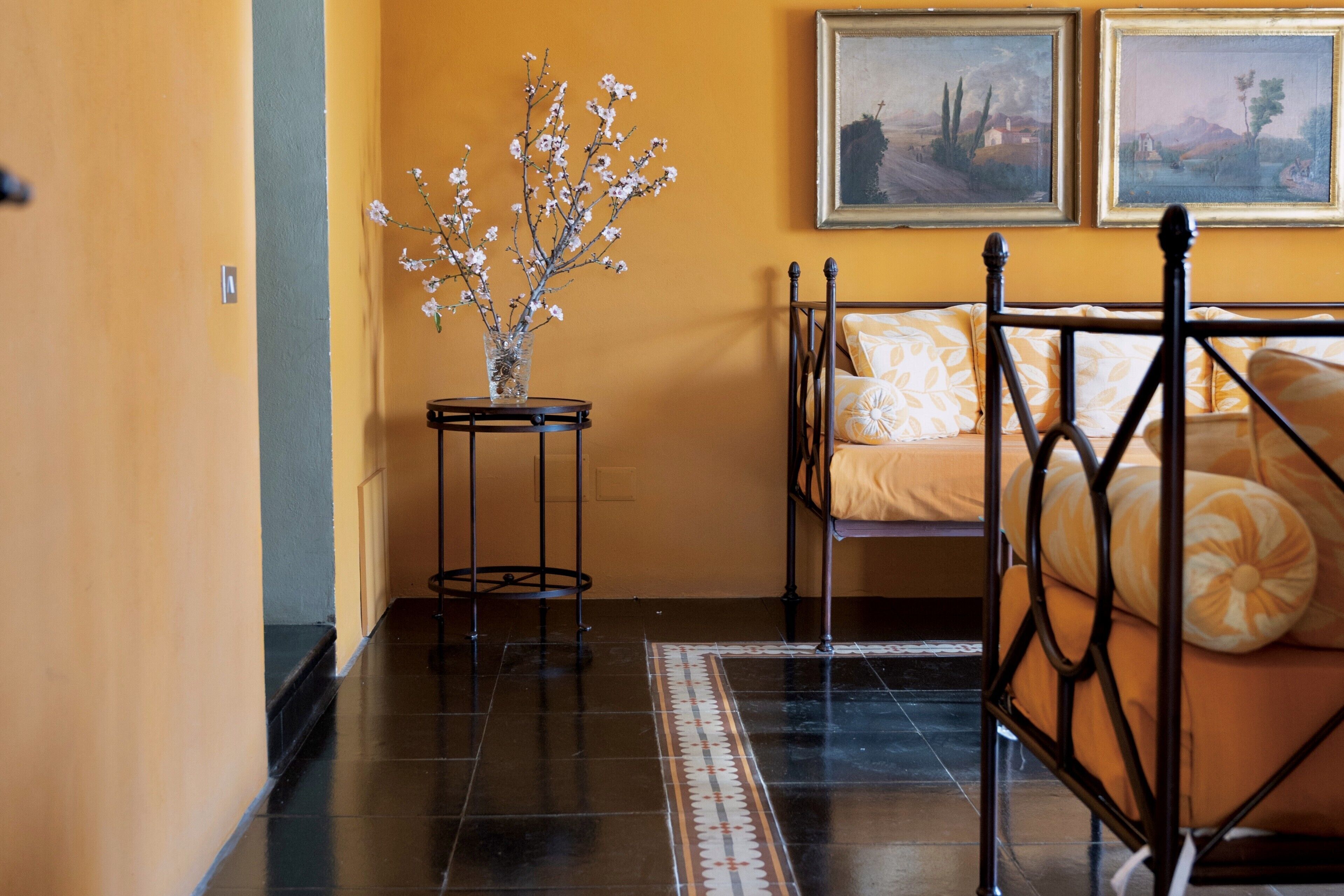 A bedroom with a metal bed frame, a bedside table with a vase of flowers, and two framed pictures on the wall.