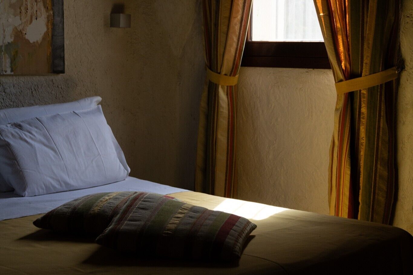 A bed with a striped pillow and a plain pillow, a window with curtains, and a wall with peeling paint.
