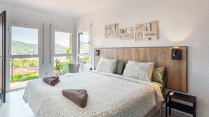 Une chambre à coucher avec un grand lit, une tête de lit en bois, une table de chevet et une vue sur la verdure par les fenêtres.