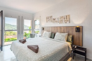 Un dormitorio con una cama grande, un cabecero de madera, una mesita de noche y vistas a zonas verdes por las ventanas.