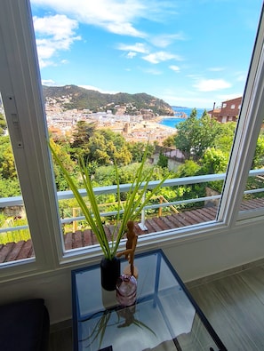 Una habitación con un ventanal grande con vistas a un pueblo costero, una mesita con un florero y una figura de madera, y una mesa de cristal.