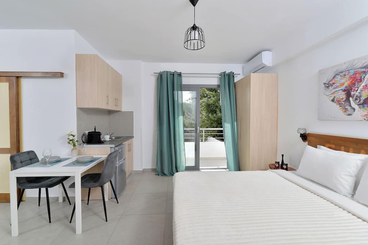 Ein modernes Hotelzimmer mit einem Bett, einem Schreibtisch mit Stühlen, einer kleinen Küchenzeile und einem Balkon mit Vorhängen.