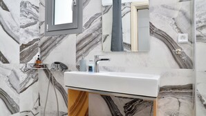 A modern bathroom with a marble backsplash, a white sink, a mirror, and a shower area. - SunDay Free Airport Shuttle (Spata-Artemida)