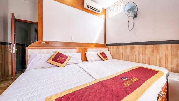 A hotel room with a wooden headboard, a bed with white bedding and a red blanket, an air conditioning unit, and a wall-mounted fan. - Hotel Airport MTT (Ho Chi Minh City)