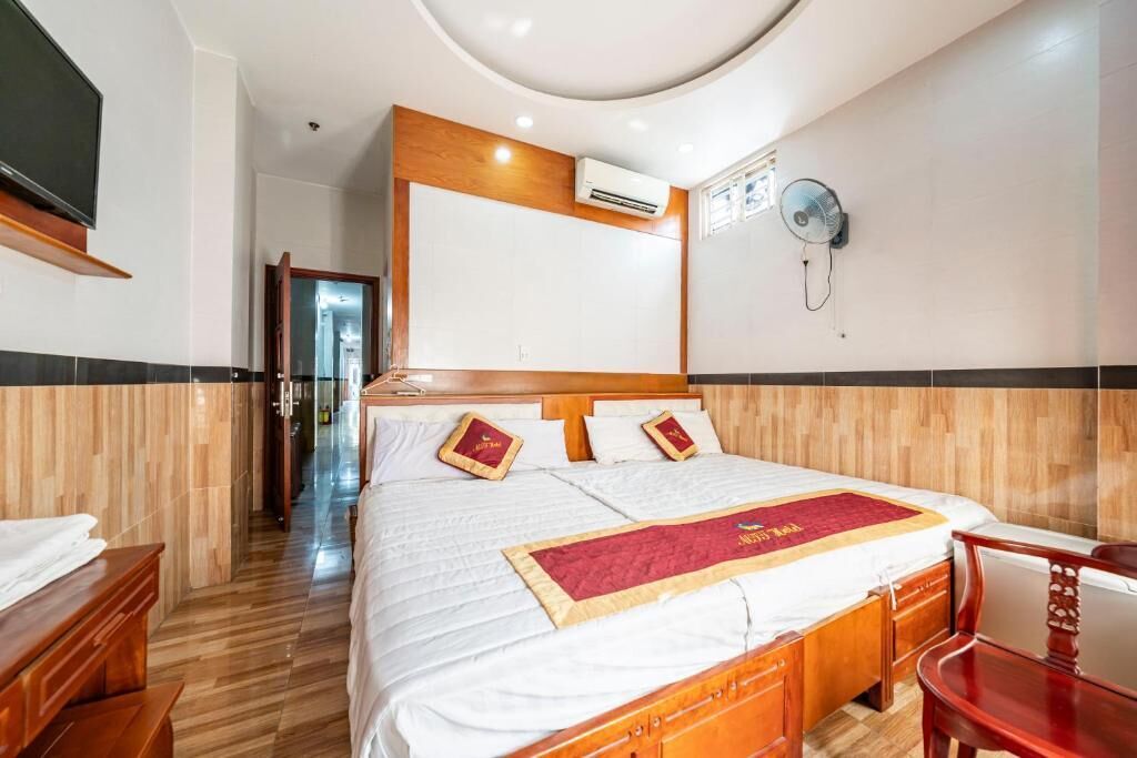 A hotel room with a bed, wooden headboard, wall-mounted air conditioning, a bedside table, a chair, and a TV.