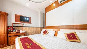 A hotel room with a large bed, a wooden headboard, a flat-screen TV mounted on the wall, and a desk with a mirror and a vase of flowers. - Hotel Airport MTT (Ho Chi Minh City)
