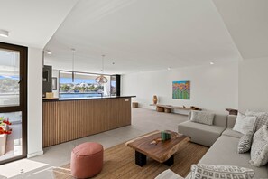 A modern living room with a sofa, a wooden coffee table, a pink ottoman, a kitchen area with a bar, and a large window with a view. - The Wharf Willemstad (Willemstad)
