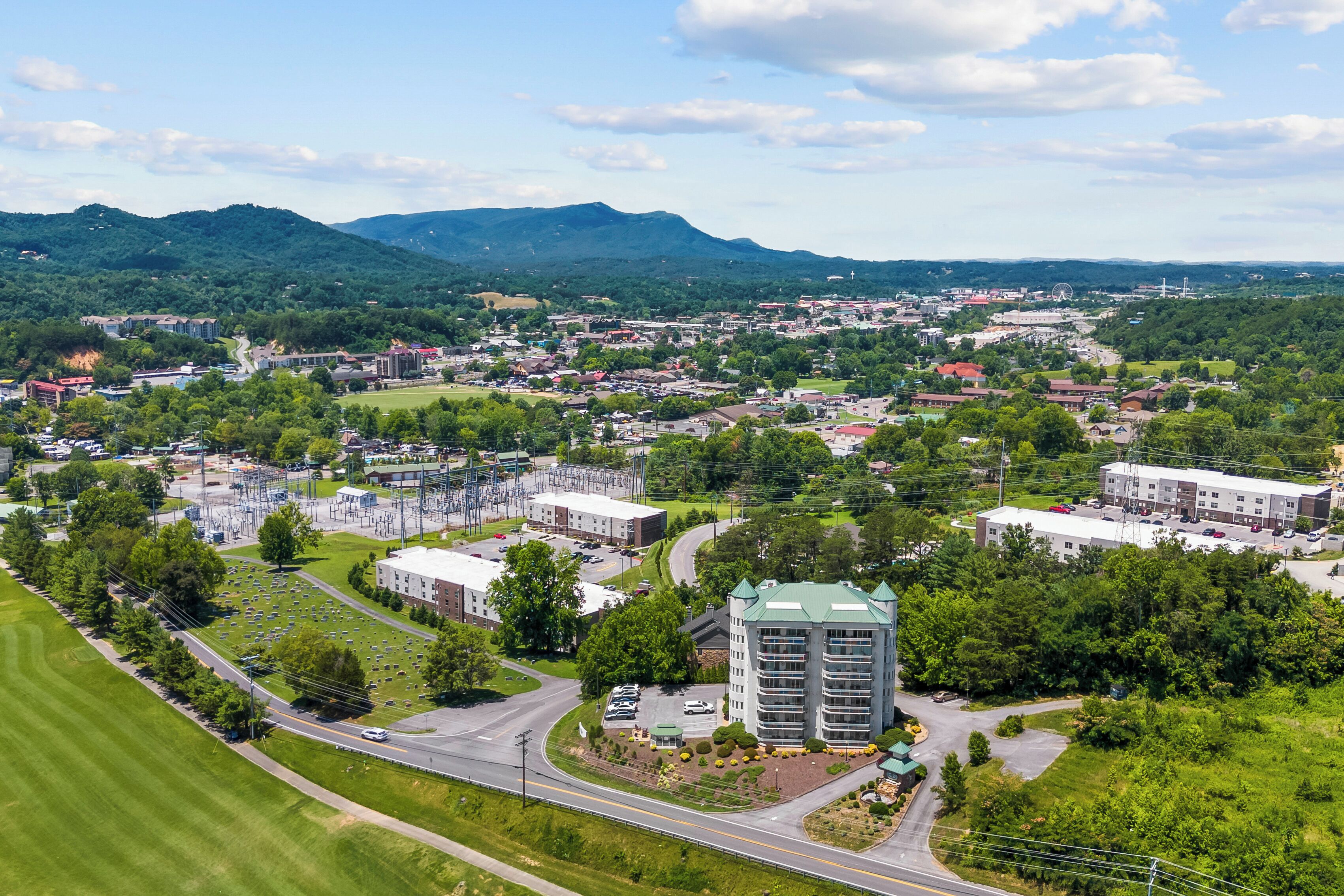 An aerial view of a town with a prominent multi-story building, surrounded by greenery and a mountain range in the background.