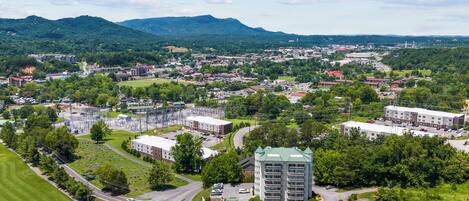 An aerial view of a town with a prominent multi-story building, surrounded by greenery and a mountain range in the background.