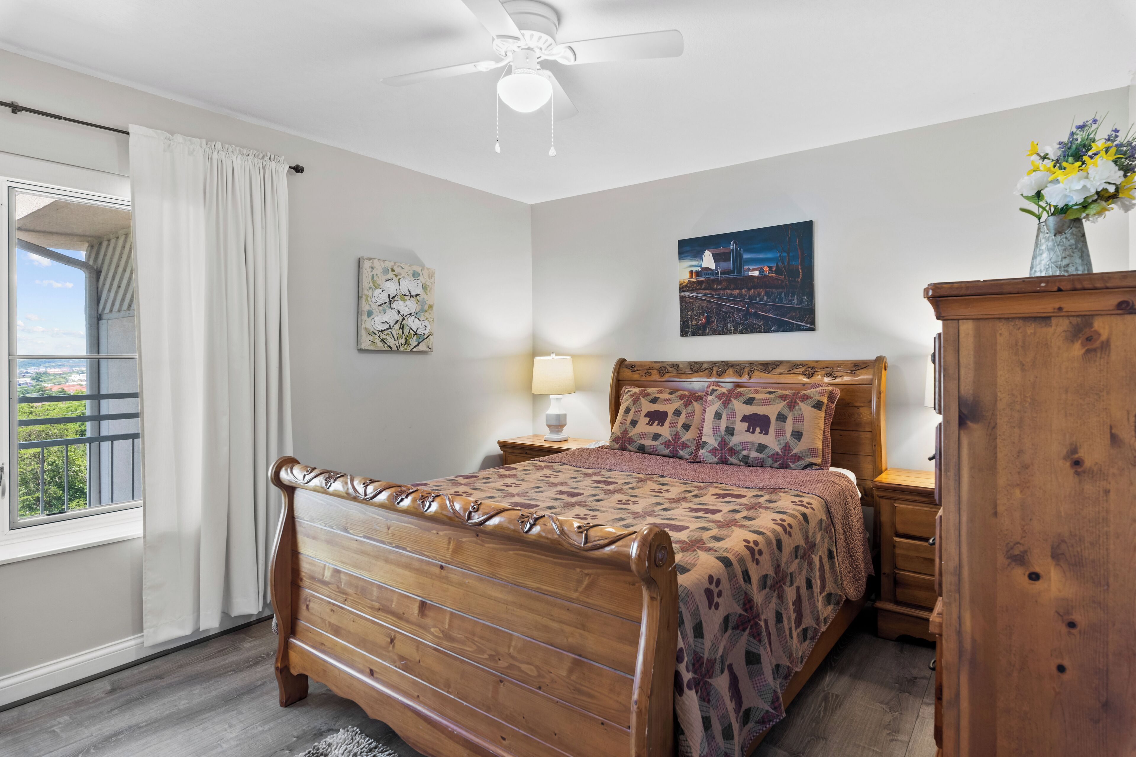 A bedroom with a wooden bed, a nightstand, a dresser, a ceiling fan, a window with curtains, and a cityscape painting on the wall.