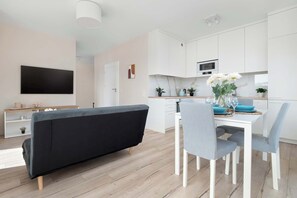 A modern living room with a black sofa, a dining table with chairs, a flat-screen TV mounted on the wall, and a kitchen area with white cabinets and a microwave. - Bright Apartment in Lodz | Parking, Balcony, Close to Museum (Łódź)
