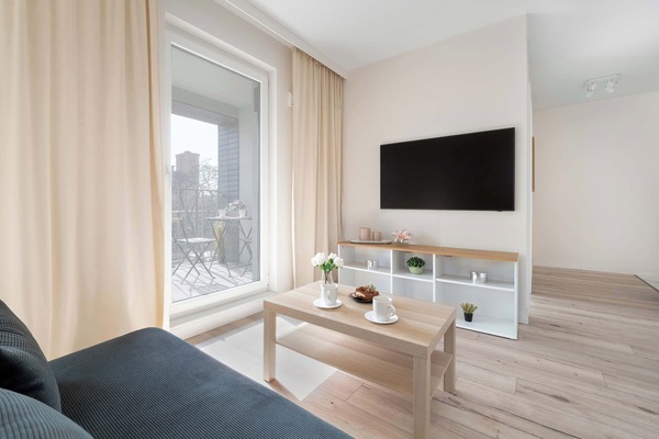 A living room corner with a television mounted on the wall. Soft natural light enters through the windows, illuminating a tidy seating area.