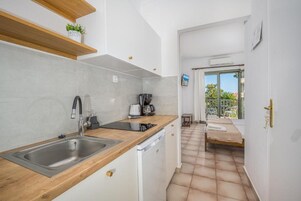 Ένας μικρός, λειτουργικός χώρος κουζίνας με νε ροχύτη, πλυντήριο πιάτων και καφετιέρα. Ένα μικρό τραπέζι με καρέκλες είναι ορατό σε μια μικρή τραπεζαρία, η οποία διακρίνεται μέσα από μια ανοιχτή πόρτα.