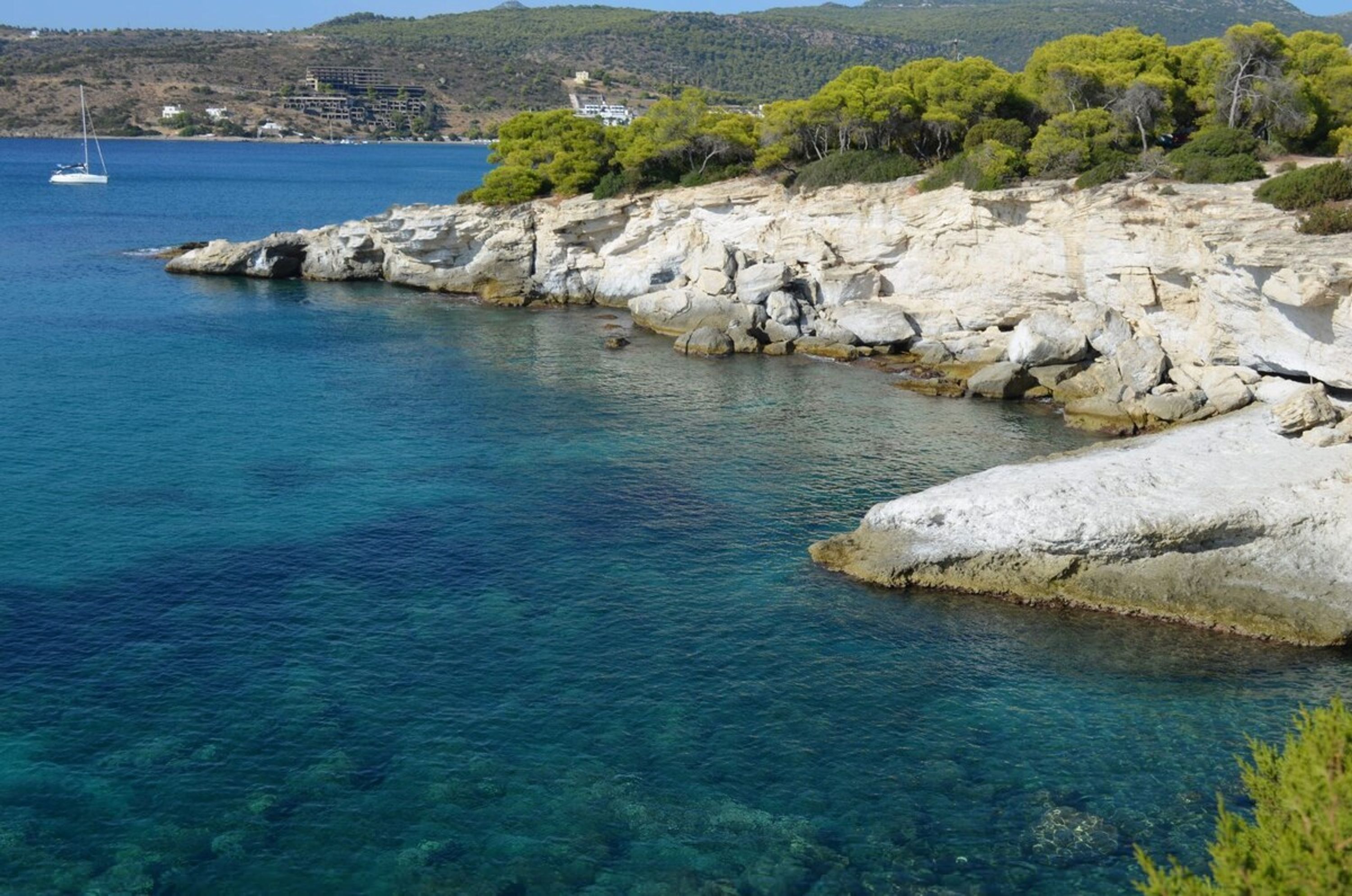 A coastal landscape with clear blue water, rocky shore, and a sailboat in the distance.