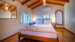 A bedroom with a wooden ceiling, a bed with white linens, a nightstand, a mirror, and a window. - My Little House - 123 Soleil (Aegina)