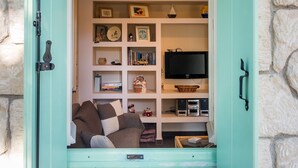 A room with a stone wall, a teal door, and a bookshelf filled with various items. - My Little House - 123 Soleil (Aegina)