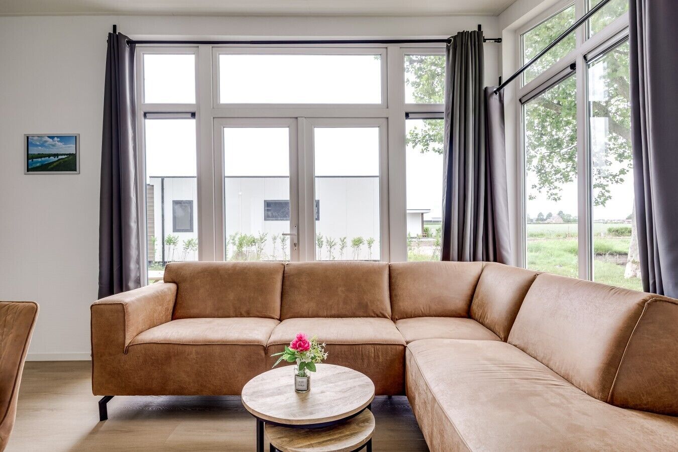 A modern living room with a large sectional sofa, a small round table with a vase of flowers, and large windows offering a view of the outdoors.