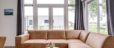 Ein modernes Wohnzimmer mit einer großen Eckcouch, einem kleinen runden Tisch mit Blumenvase und großen Fenstern mit Blick ins Freie.