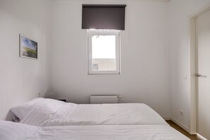 Une chambre avec deux lits, une fenêtre avec un store, un radiateur et une porte.