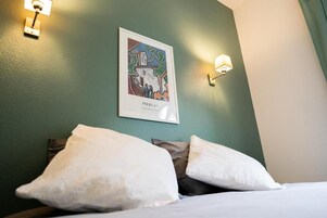Quarto de hotel com cama, almofadas, uma moldura na parede e luminárias fixadas na parede.
