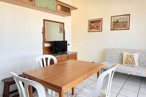 Una habitación con una mesa de madera, sillas blancas, un televisor, un sofá con un cojín floral y dos cuadros en la pared.