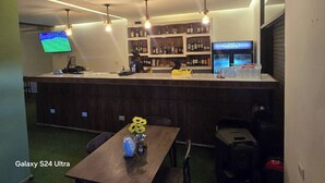 Un espace bar avec un comptoir, des étagères remplies de bouteilles, un téléviseur et une table ornée d’un vase de tournesols.