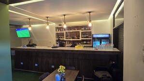 Un espace bar avec un comptoir, des étagères remplies de bouteilles d’alcool, un téléviseur et une table agrémentée d’un vase.