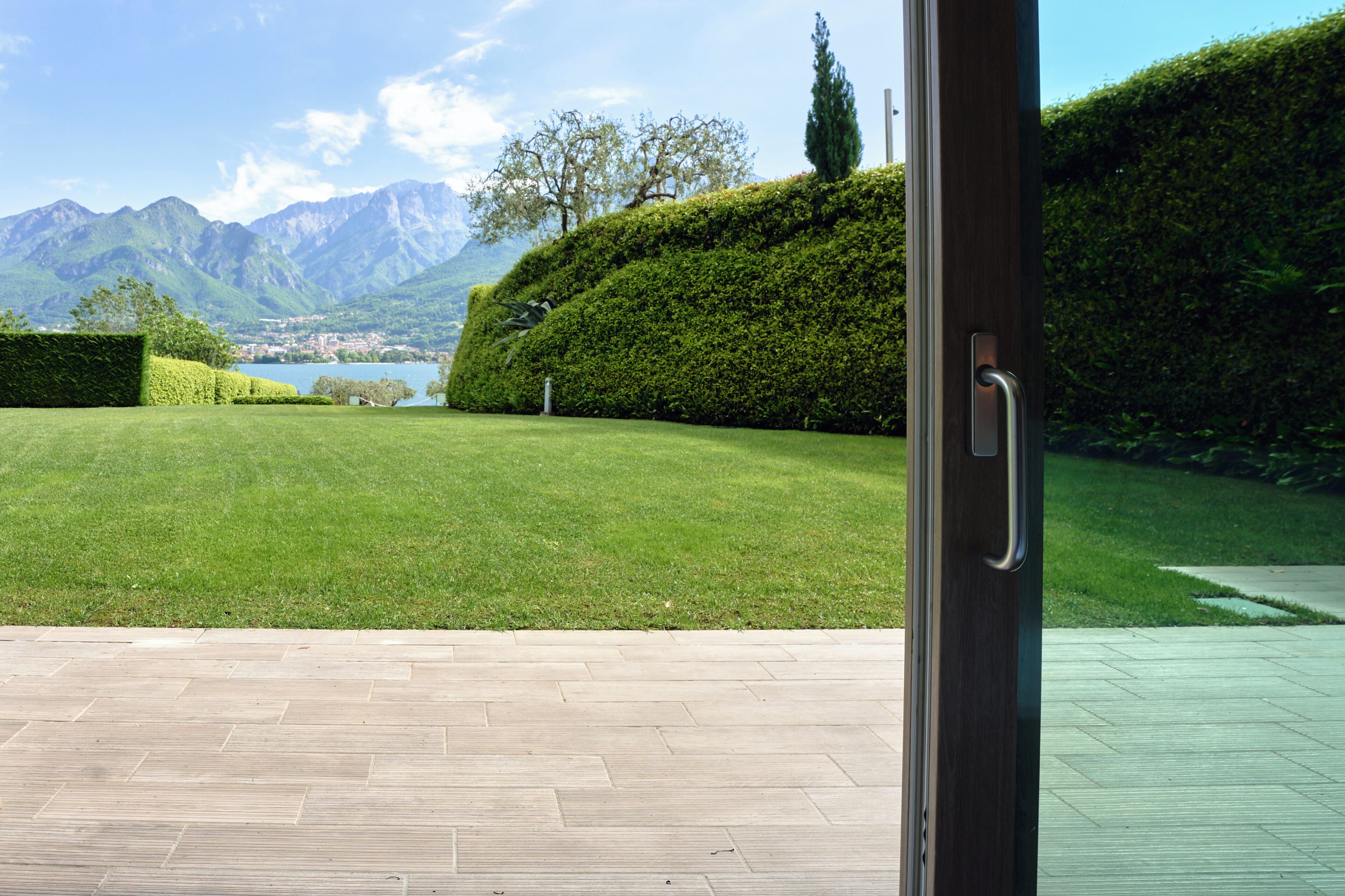 A view from a room with a large glass door opening to a well-maintained lawn, a hedge, and a mountain backdrop.