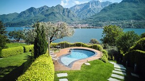 A pool with a wooden deck, surrounded by well-manicured hedges and trees, with a mountainous backdrop. - Villa Costanza (Oliveto Lario)