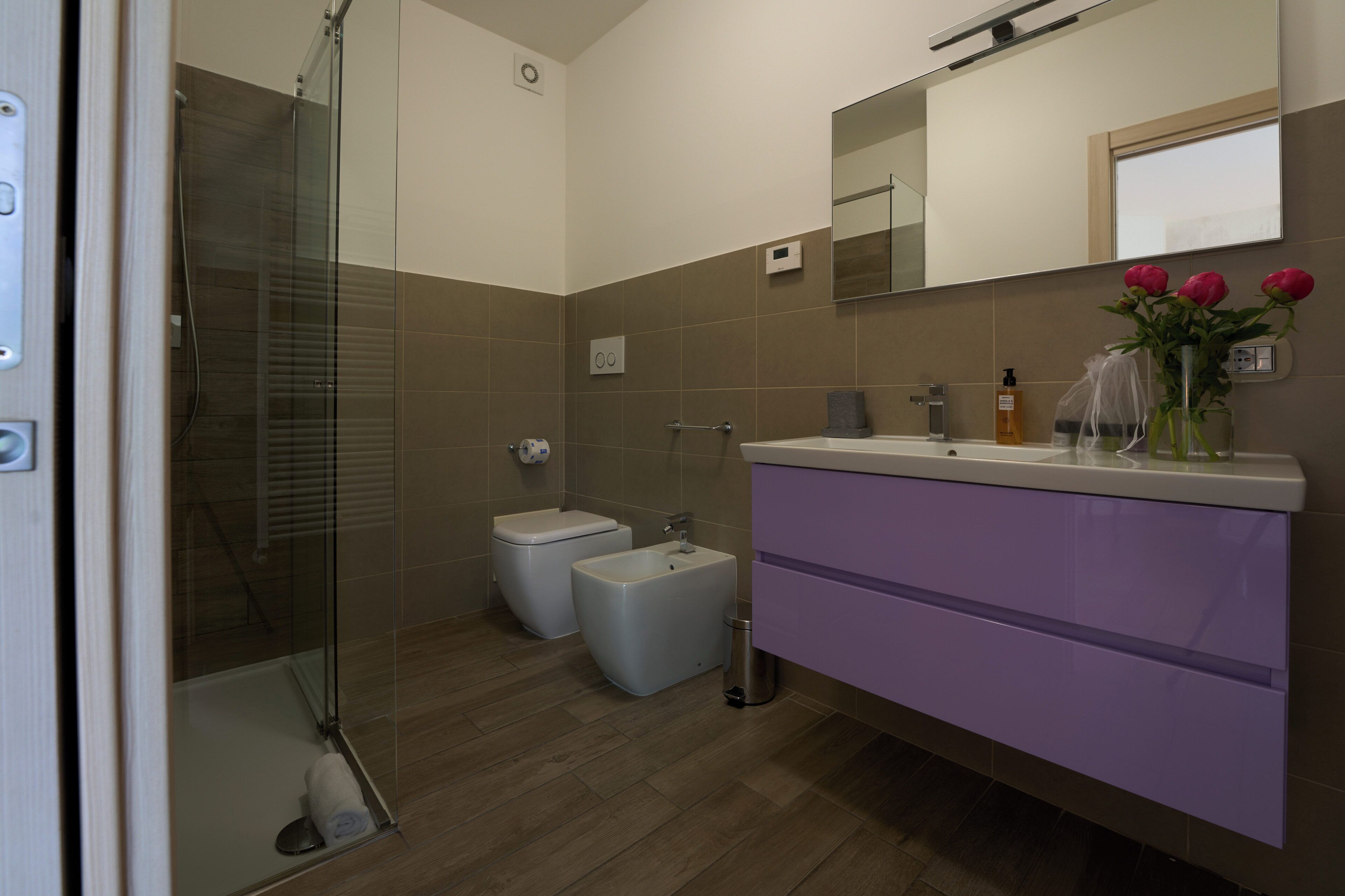 A modern bathroom with a glass shower enclosure, two toilets, a vanity with a mirror, and a vase with flowers on the counter.