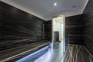 Un baño moderno con paredes y suelo de mármol negro, cabina de ducha de vidrio y un banco.