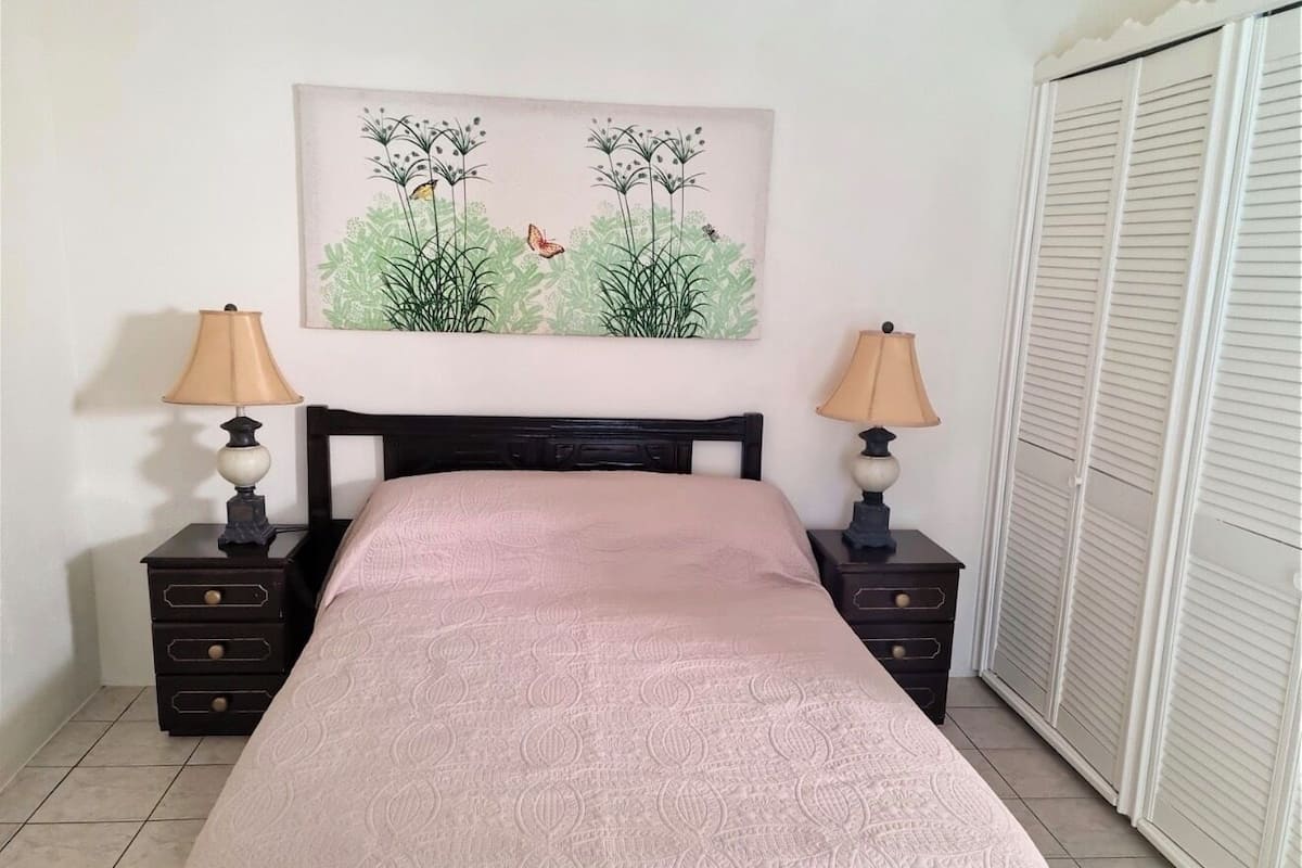 A bedroom with a bed, two nightstands, a lamp on each, a painting above the bed, and a wardrobe to the right.