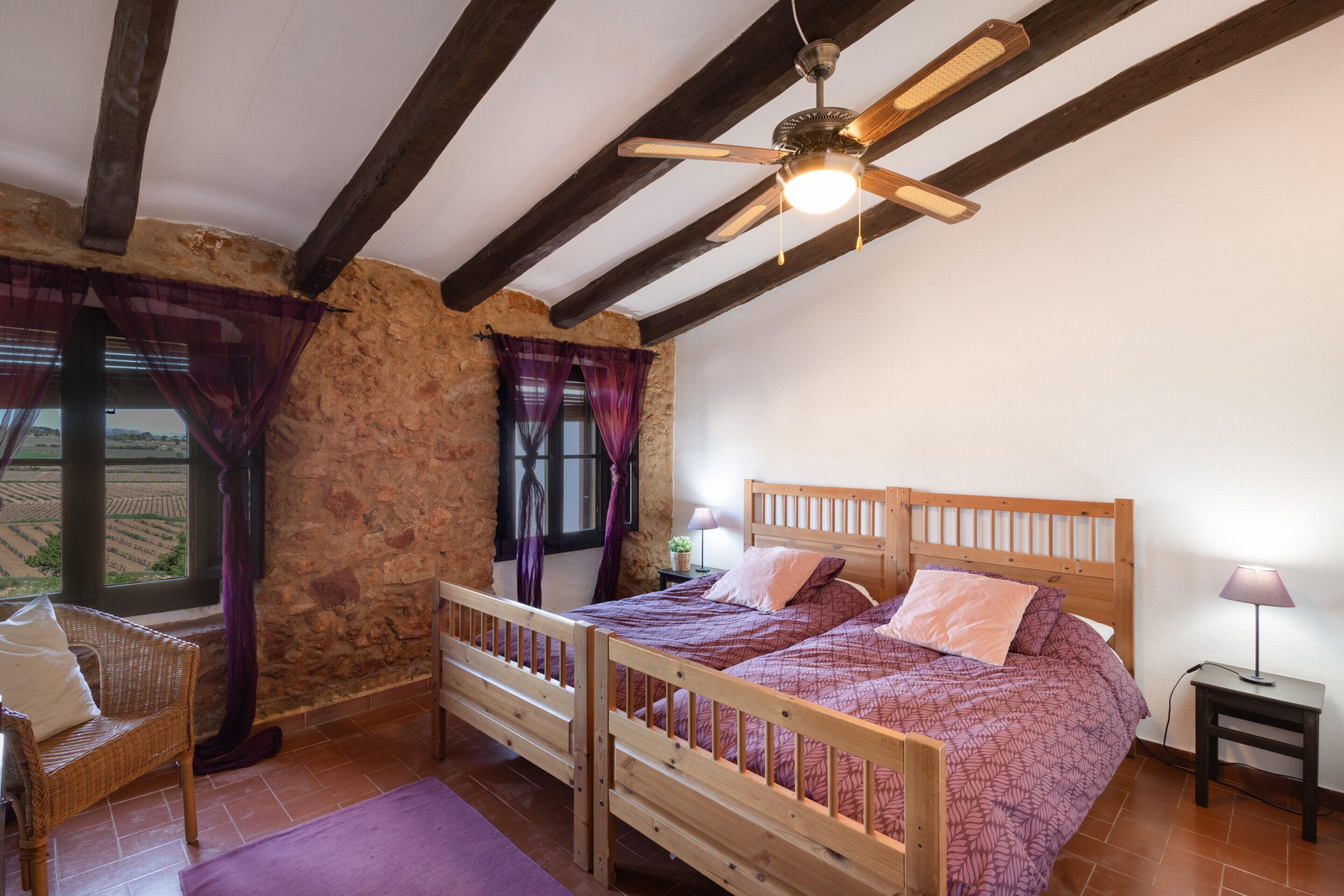 Une chambre à coucher avec un lit en bois, un mur en pierre, une fenêtre avec des rideaux et un ventilateur de plafond.