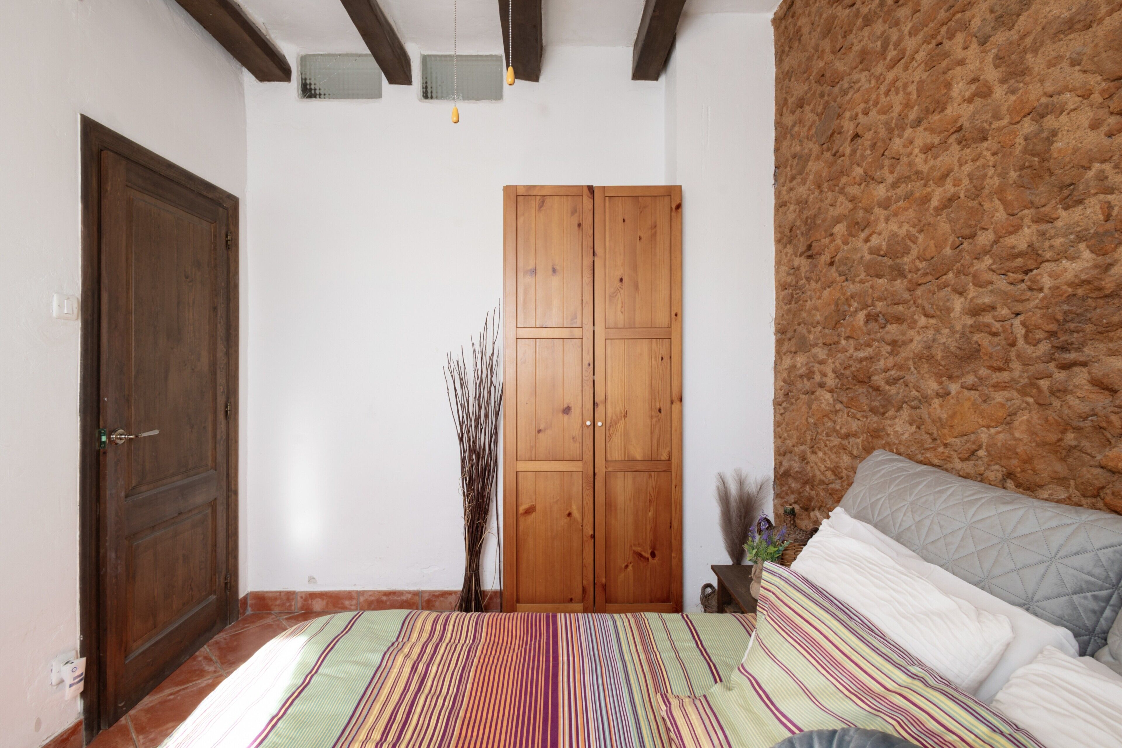 Un dormitorio con cama, un armario de madera, pared de piedra y suelo de baldosas.