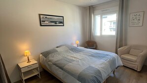 Un dormitorio con cama, una silla, mesita de noche, ventilador de techo, ventana y un cuadro colgado en la pared.