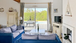 Un salon avec un canapé, une table, un téléviseur et une vue sur une piscine et un espace verdoyant.