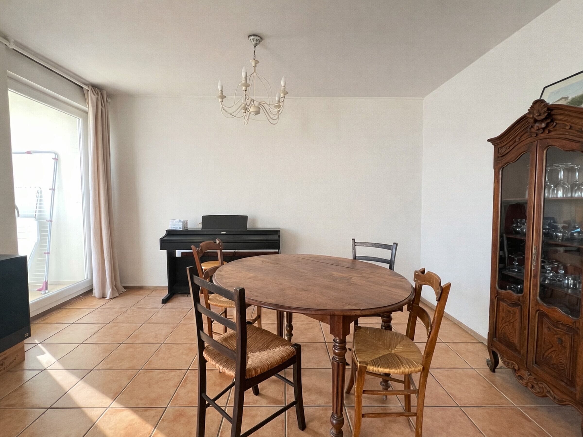 Un comedor con una mesa redonda de madera, cuatro sillas, una lámpara de araña, un piano y un aparador con puertas de cristal.