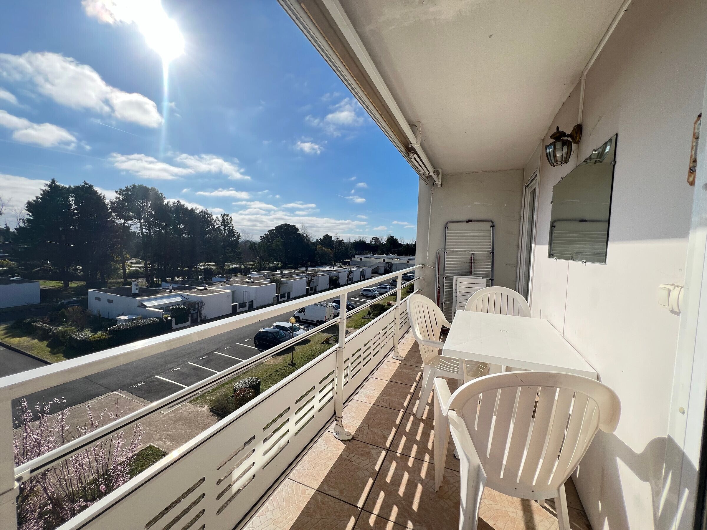 A balcony with a view of a parking lot and buildings, white chairs, and a table.