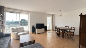 A spacious living area with a sofa, a coffee table, a dining table with chairs, a television, and a bookshelf. - Appartement lumineux avec terrasse – Arcachon, Ville d’Automne (Arcachon)