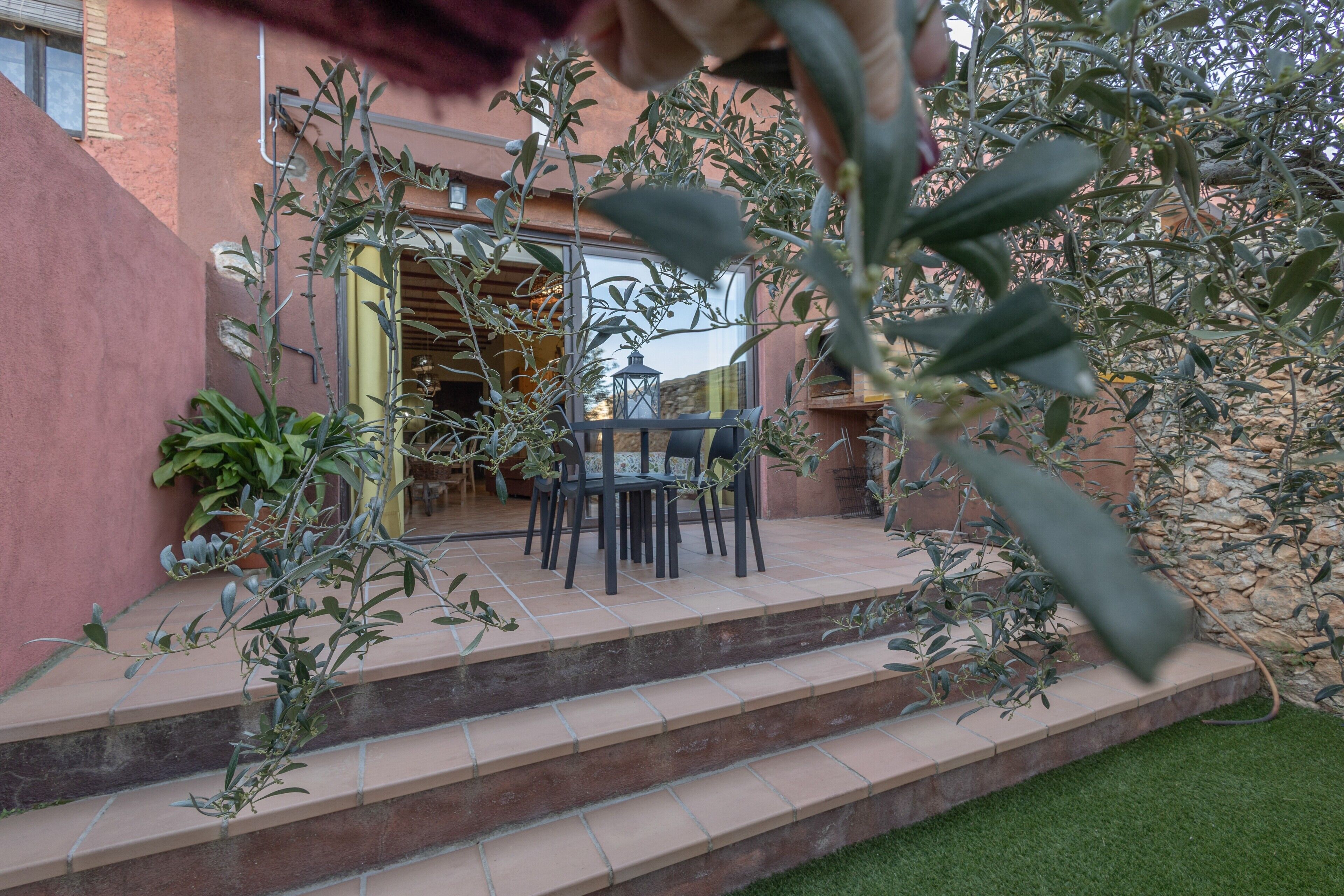 Un patio con mobiliario exterior, escalones que llevan a la entrada de un edificio y vegetación circundante.