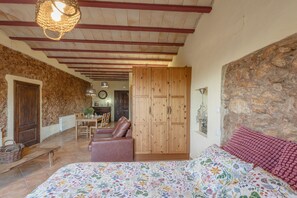 Ein Zimmer mit einem Bett, einem Holzschrank, einer Steinwand, einer Holzbalkendecke und einem Essbereich mit Tisch und Stühlen.