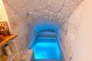 Quarto com paredes de pedra, uma banheira iluminada em azul, uma prateleira de madeira com objetos e um recipiente branco.