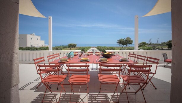An outdoor dining area with a table set for six, surrounded by red chairs, overlooking a scenic view of the sea and a building. - Exclusive beachfront villa, 6 bedrooms, garden, Monopoli (Monopoli)