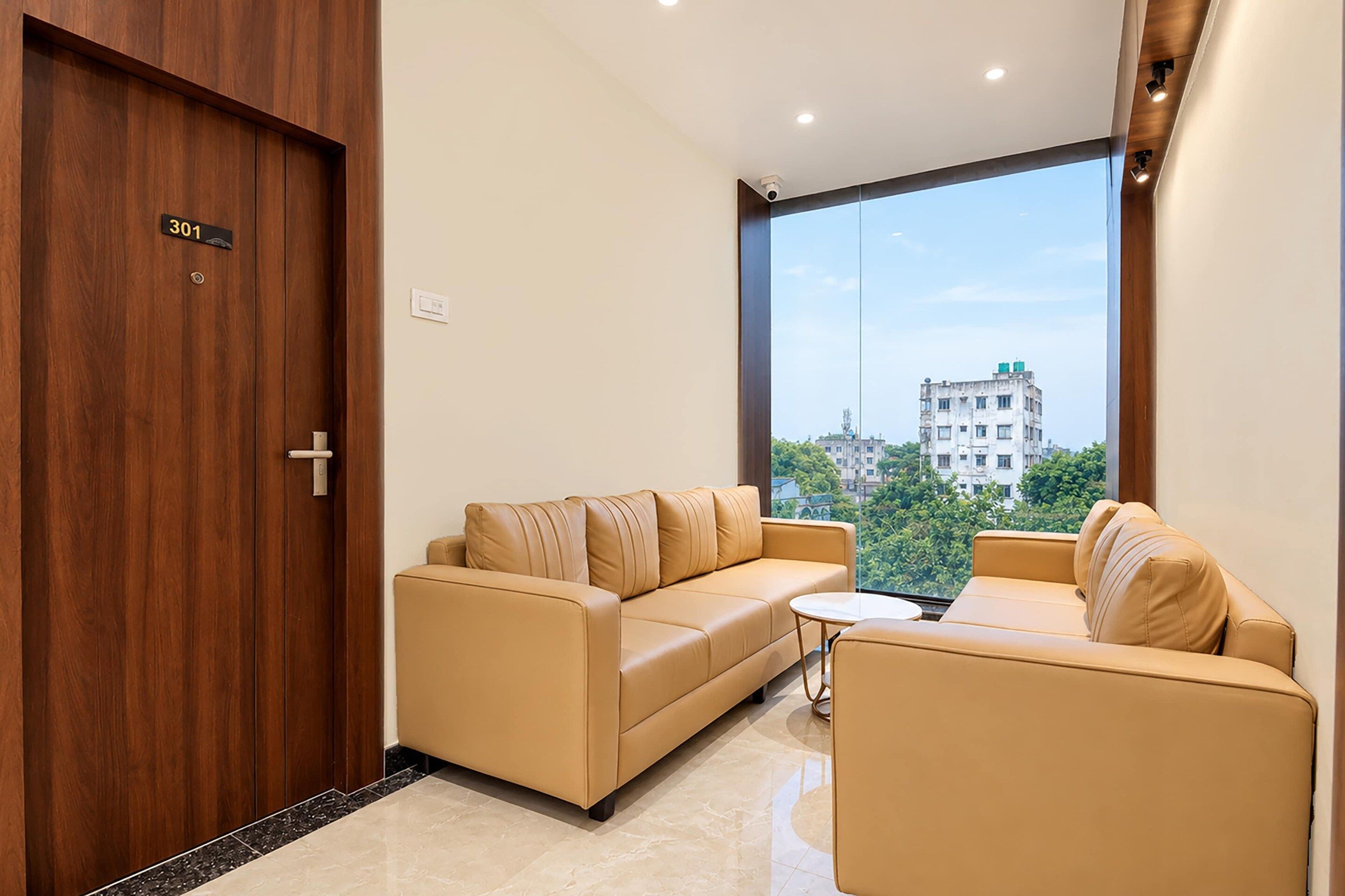 A modern living room with a sofa and armchair, a small table, a wooden door with a number plate, and a large window offering a city view.